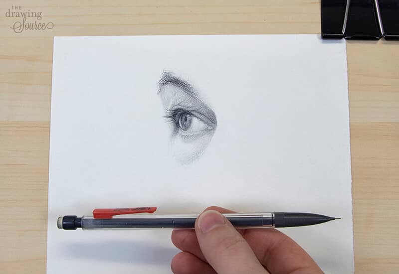 A realistic eye drawn with a single mechanical pencil, held up beside the finished drawing — showing that beautiful, realistic drawing needs only simple materials.